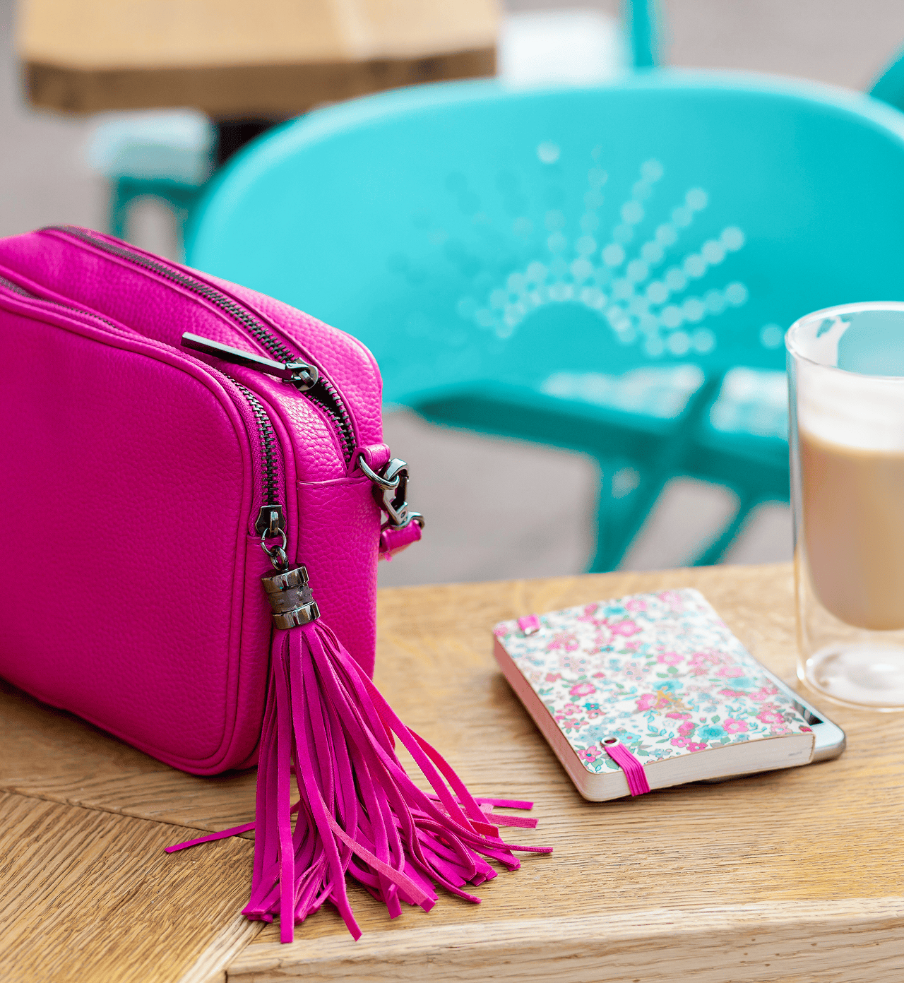 Bright pink leather crossbody bag with tassel placed on a wooden café table beside a floral notebook and latte
