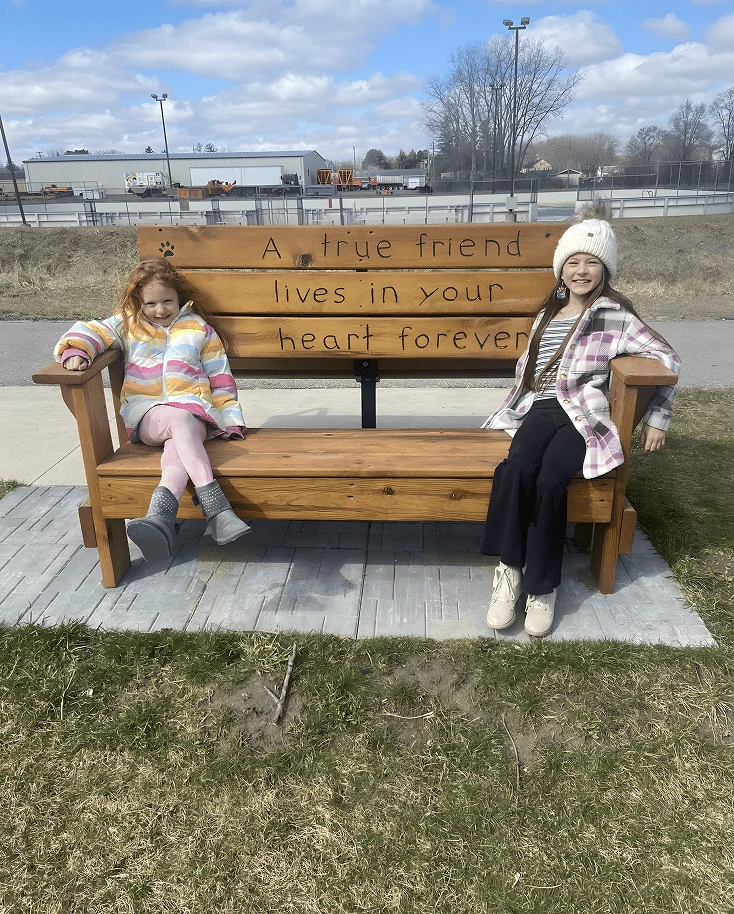 Cassie Large’s daughters sitting together on a large wooden friendship bench engraved with “A true friend lives in your heart forever,” enjoying a chilly day in Bad Axe, Michigan