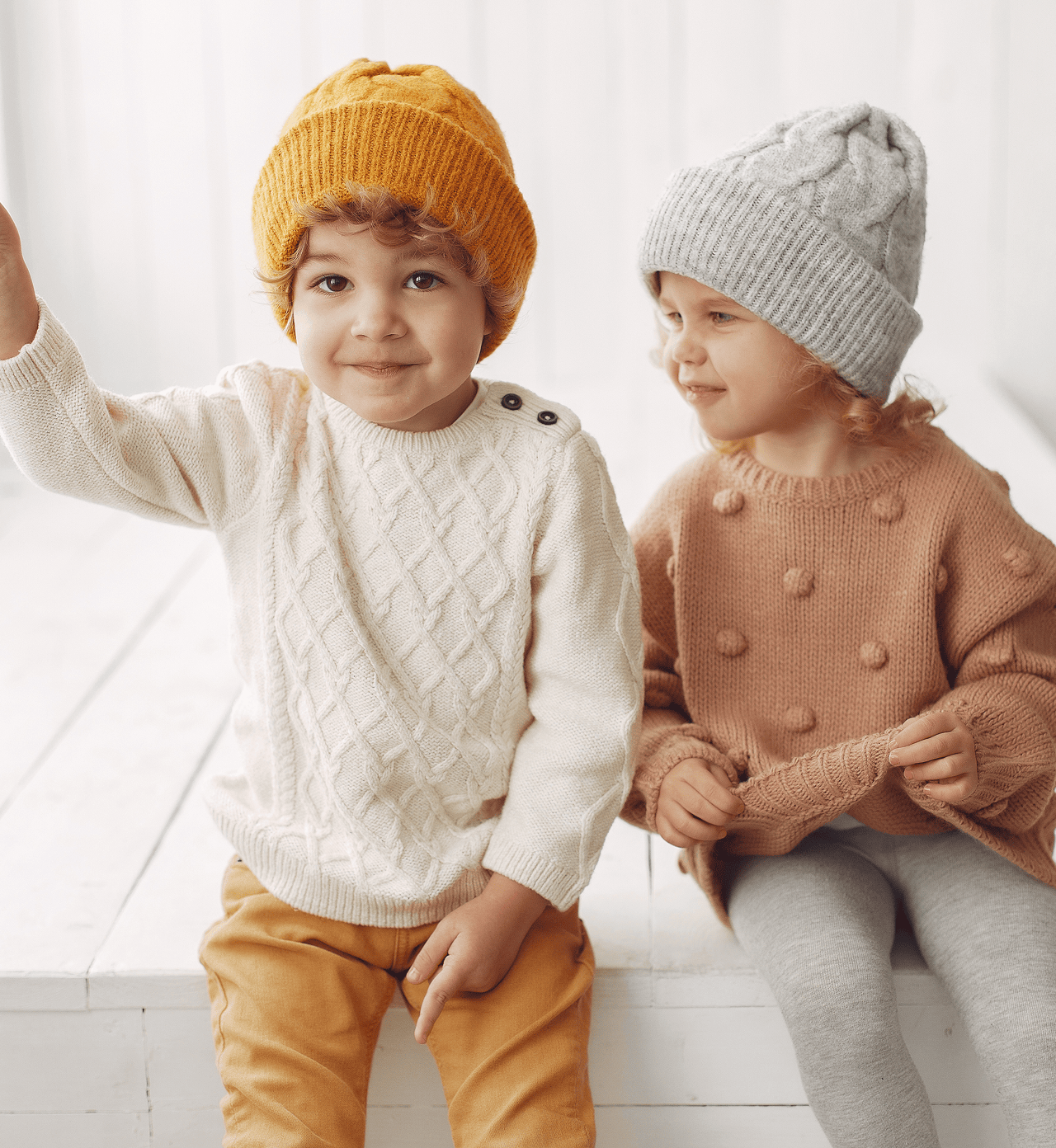 Two young children sitting together wearing cozy knit sweaters and beanies in neutral tones 